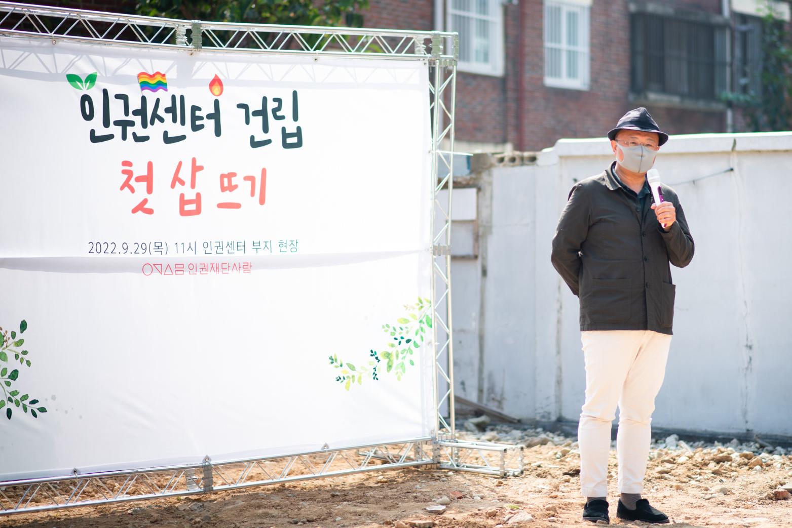 중절모와 마스크를 착용한 한 남성이 마이크를 들고 발언하고 있다. 뒤 쪽 무대에는 인권센터 건립 첫 삽 뜨기라는 문구가 보인다.