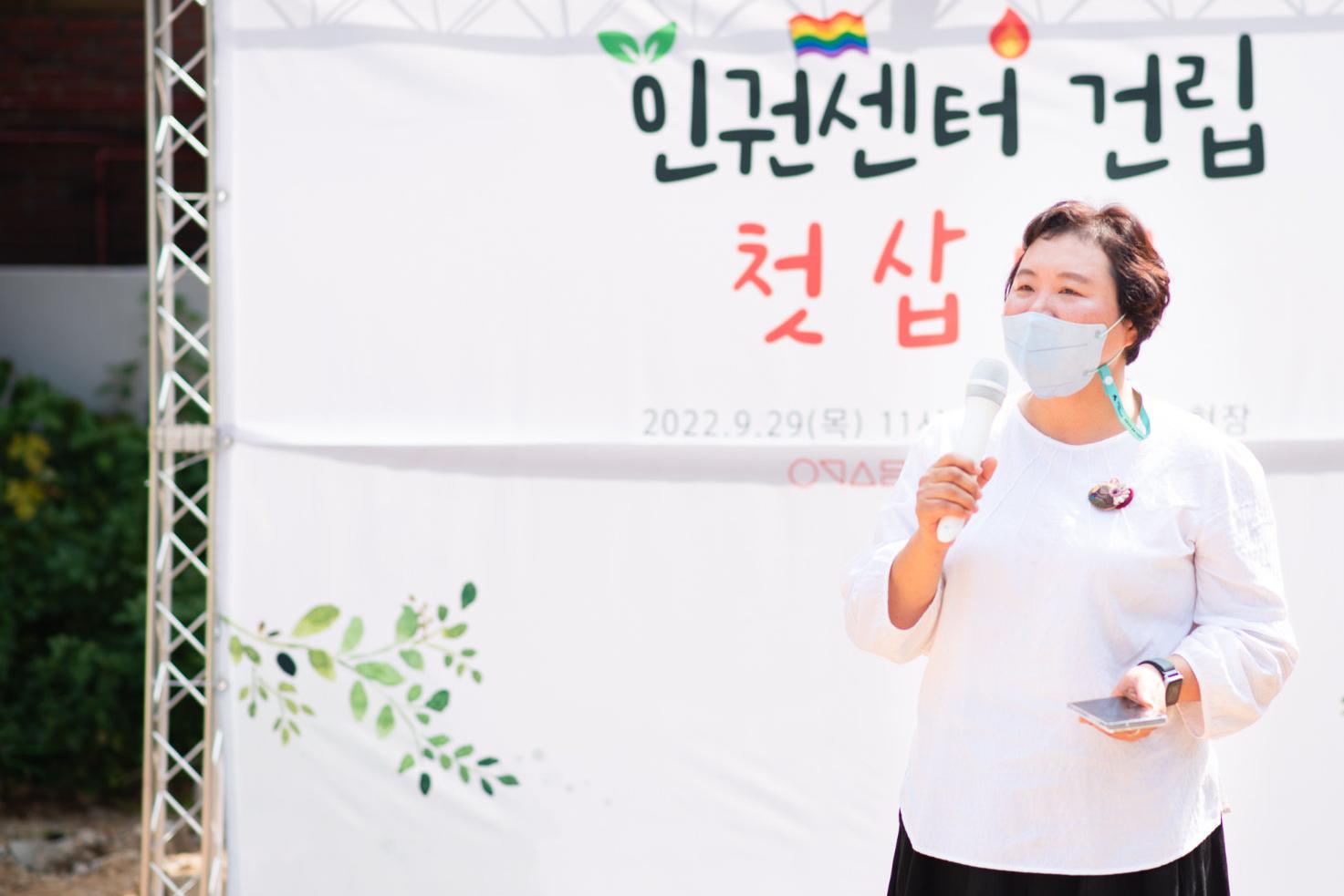 흰 티셔츠를 입고 마스크를 착용한 한 여성이 마이크를 들고 발언하고 있다.