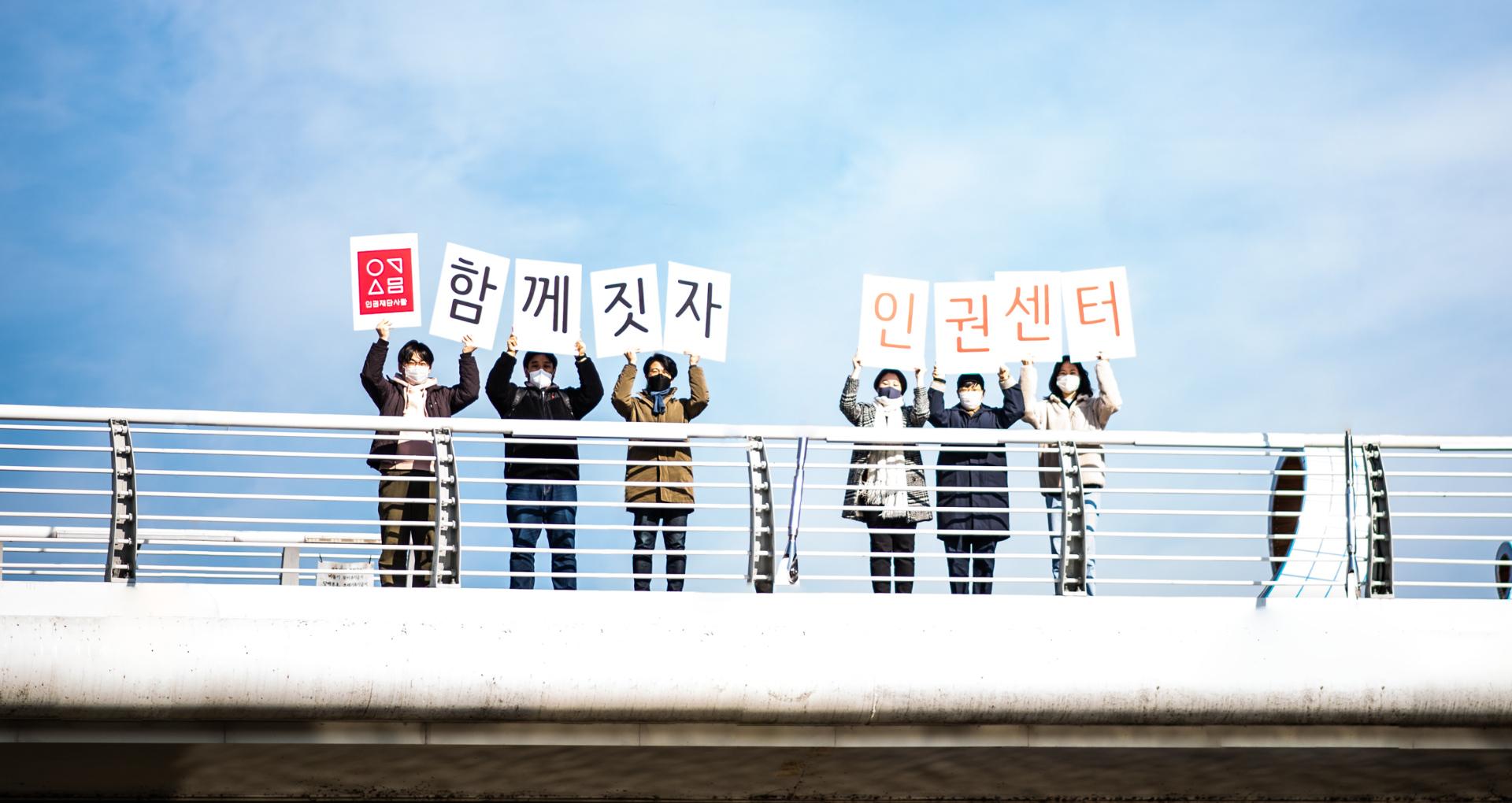 인권재단의 활동가 6명이 다리 위에서 '함께짓자 인권센터'라고 쓰인 손팻말을 들고 있다